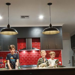 Two boys are in a kitchen with red tiled splashback and dark grey cabinets, illuminated by two black pendant lights