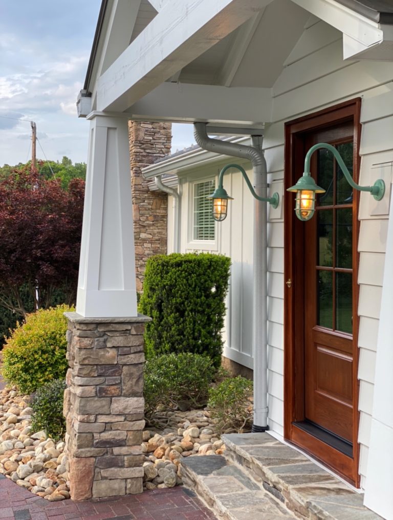 Two green gooseneck wall lights illuminate the entrance of a home with a stone pillar and wooden door