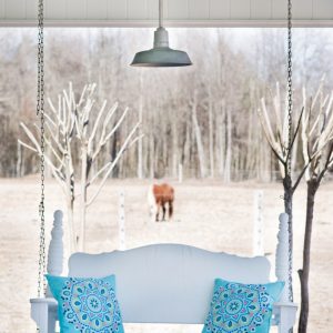 A light grey barn-style pendant light hangs above a white porch swing with turquoise cushions, overlooking a field with a horse
