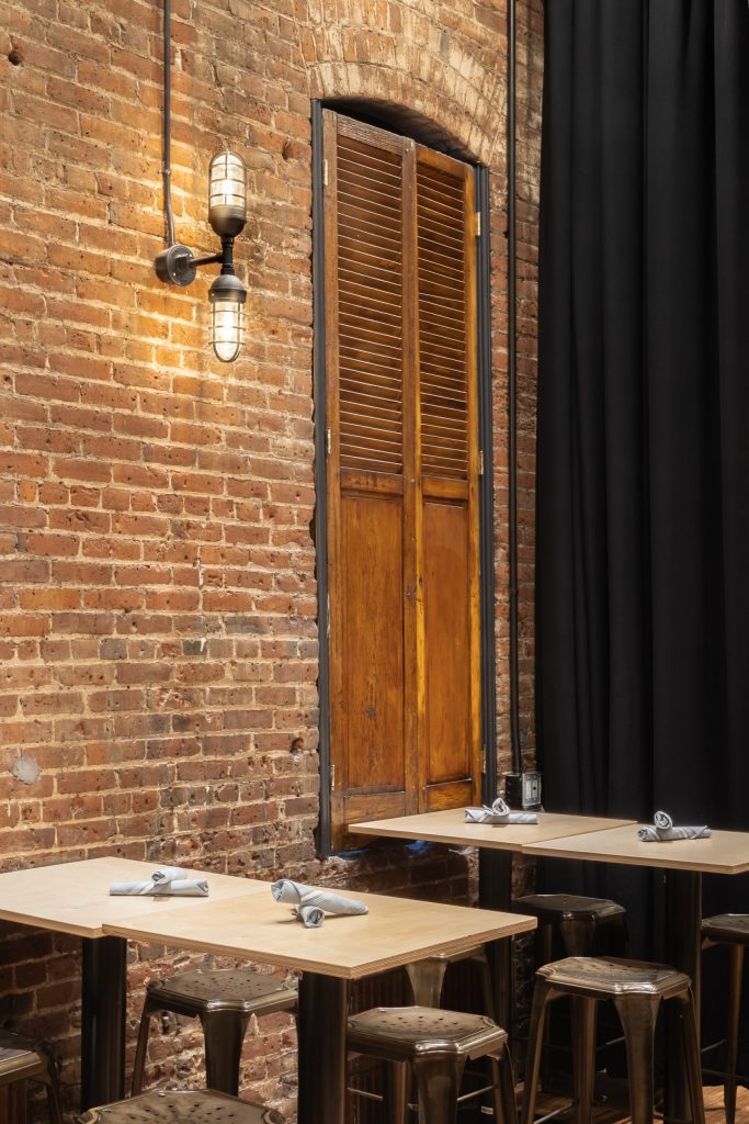 A double-headed industrial wall light illuminates a brick wall next to a wooden shuttered window in a restaurant setting