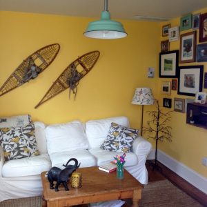 A bright yellow living room features a white sofa, a wooden coffee table, and a mint green pendant light hanging from the ceiling