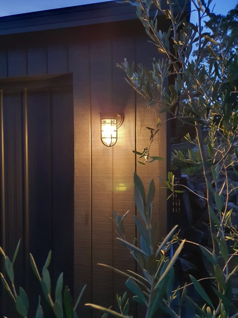 A caged outdoor wall light illuminates a dark wooden wall at dusk