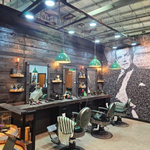 A barber shop interior features green pendant lights hanging above barber chairs and a wooden counter