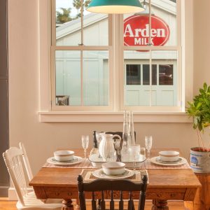 A turquoise pendant light hangs above a wooden dining table set for a meal, with a vintage Arden Milk sign visible through the window