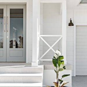A modern black gooseneck wall light is mounted on the white weatherboard exterior of a home, next to a double glass door and a garage