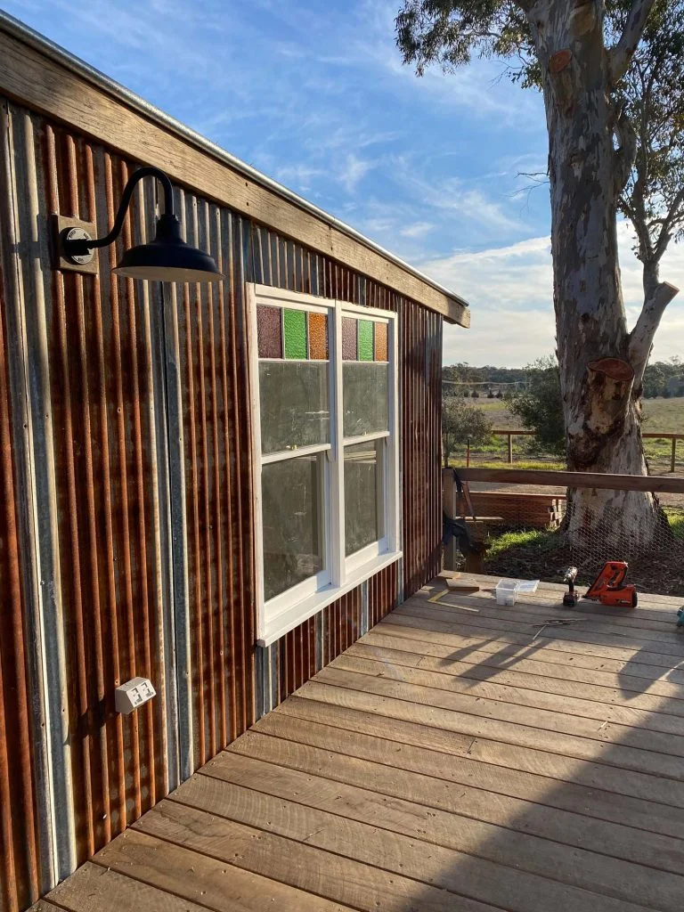 A black gooseneck wall light is mounted on a rustic corrugated iron wall next to a window with stained glass