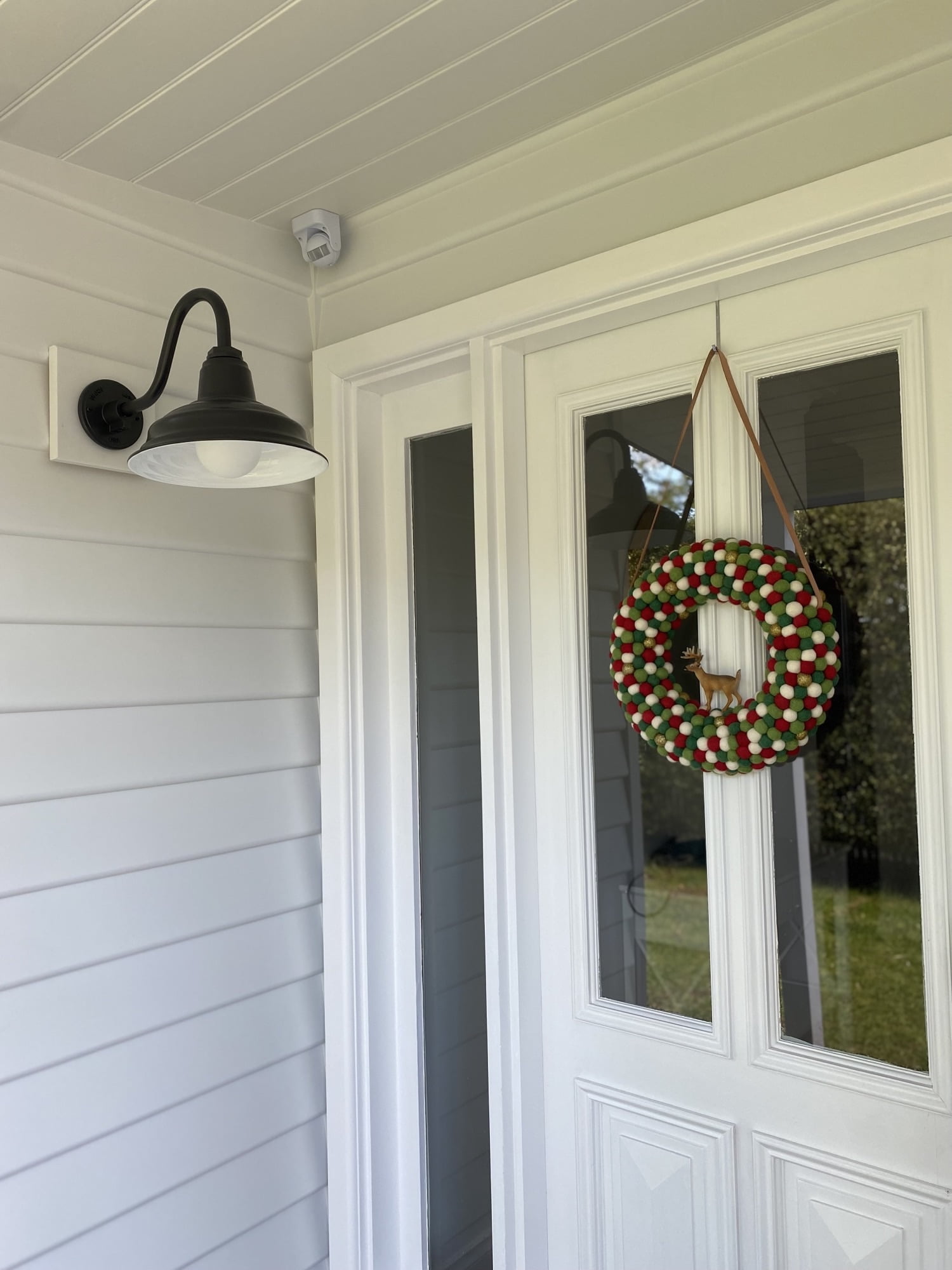 A black gooseneck wall light is mounted next to a white front door adorned with a festive wreath