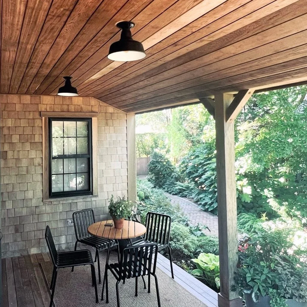 Two black barn-style pendant lights illuminate a covered porch with a wooden ceiling and shingled walls