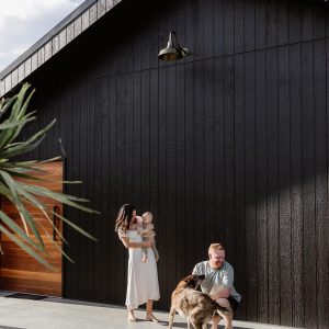 A family with their dog poses in front of a modern black timber-clad home with a gooseneck wall light