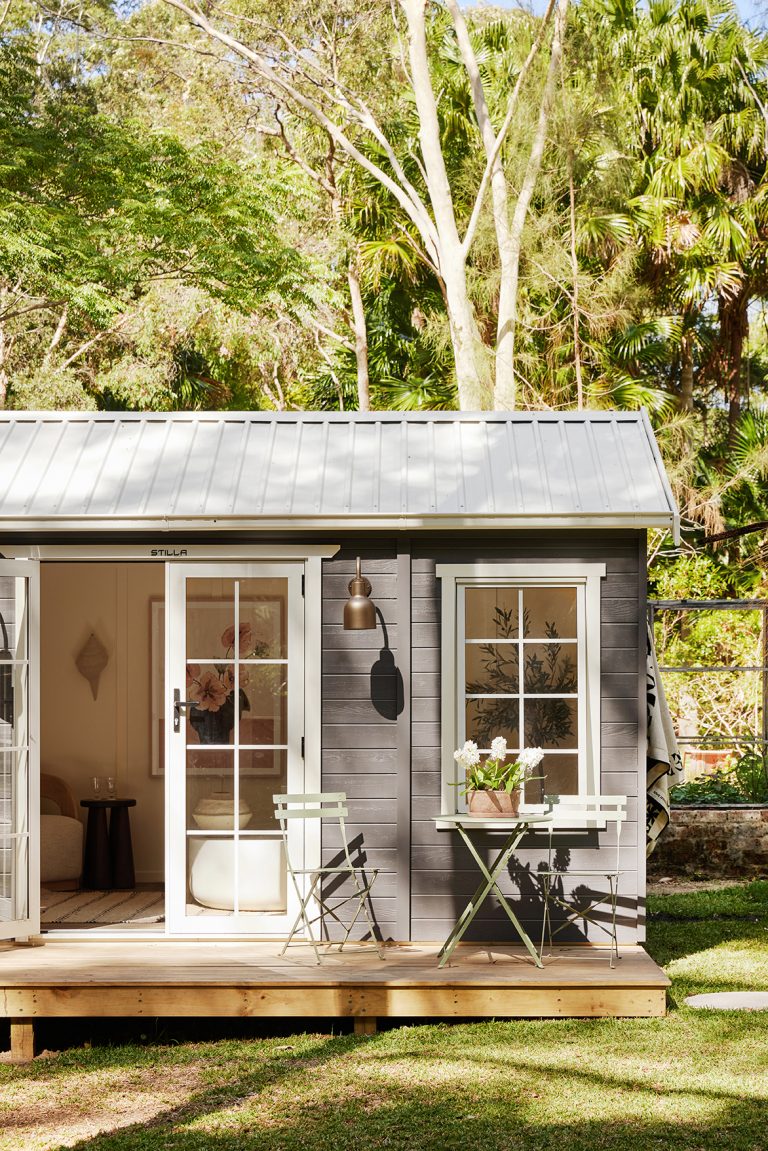 A dark grey wooden shed with a metal roof is set amongst lush greenery, featuring a wall-mounted light fixture and a small outdoor seating area