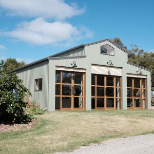 A corrugated iron shed with large wooden-framed glass doors is illuminated by three gooseneck wall lights under a bright blue sky