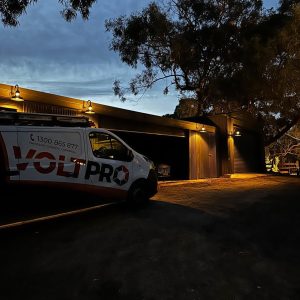 Outdoor wall lights illuminate a corrugated iron shed at dusk, with a VoltPro service van parked in front