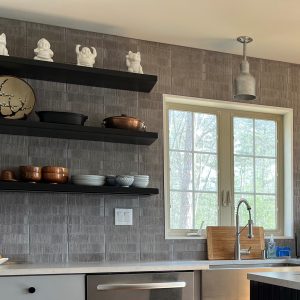 A modern kitchen features a grey tiled backsplash, open shelving with decorative items, and a pendant light fixture above a sink