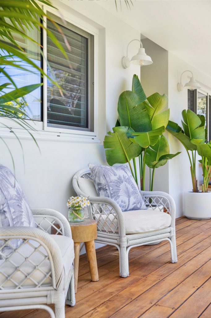 Two white wall lights illuminate a covered outdoor patio with white wicker chairs and lush green plants