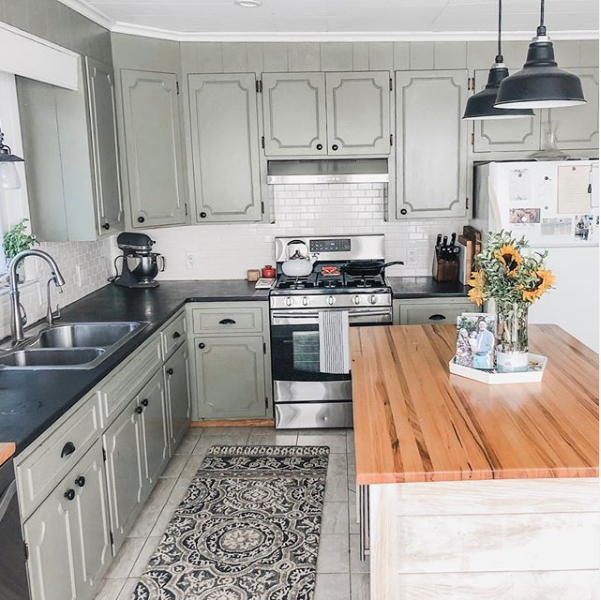 A modern farmhouse kitchen features sage green cabinetry and black pendant lights hanging over a wooden island