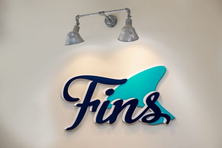 A double-headed industrial-style wall light illuminates a sign with the word "Fins" and a turquoise shark fin