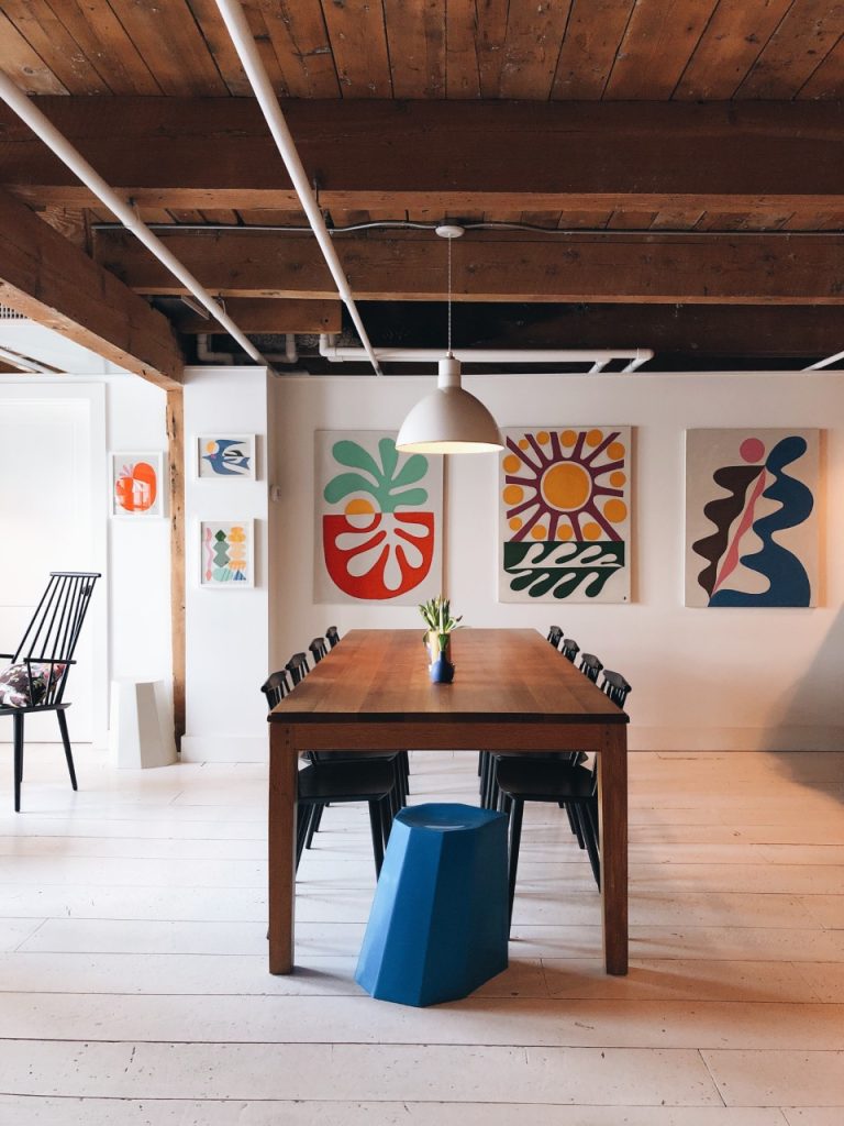 A modern dining room features a long wooden table with chairs, a blue stool, and colourful abstract art on the wall