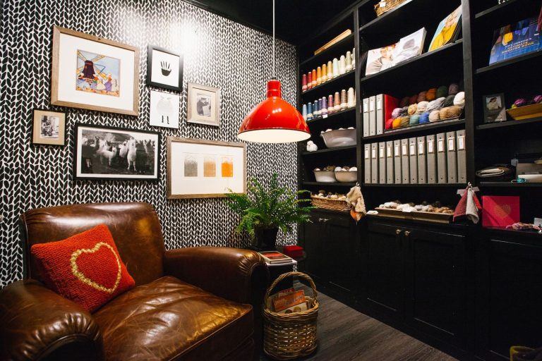 A red pendant light hangs above a brown leather armchair in a room with black and white patterned wallpaper and black shelving filled with craft supplies