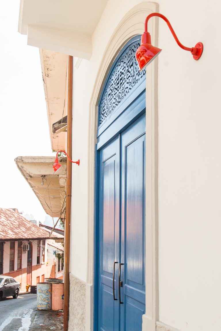 Two bright red gooseneck wall lights are mounted on a white exterior wall next to a large blue double door