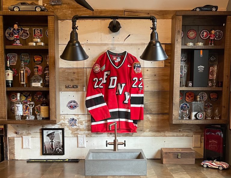A red hockey jersey hangs on a wooden wall above a concrete sink, flanked by shelves displaying liquor bottles and memorabilia
