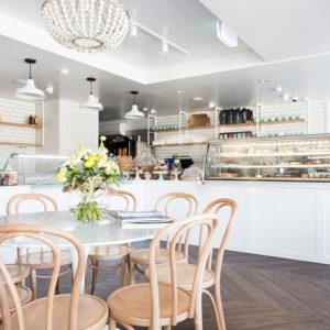 A bright and airy cafe interior features a central dining table surrounded by bentwood chairs, with pendant lights hanging from the ceiling