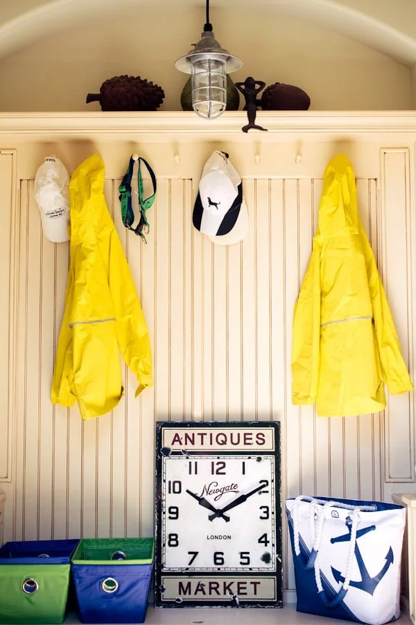 A collection of yellow raincoats and hats hang on hooks above storage bins and an antique clock