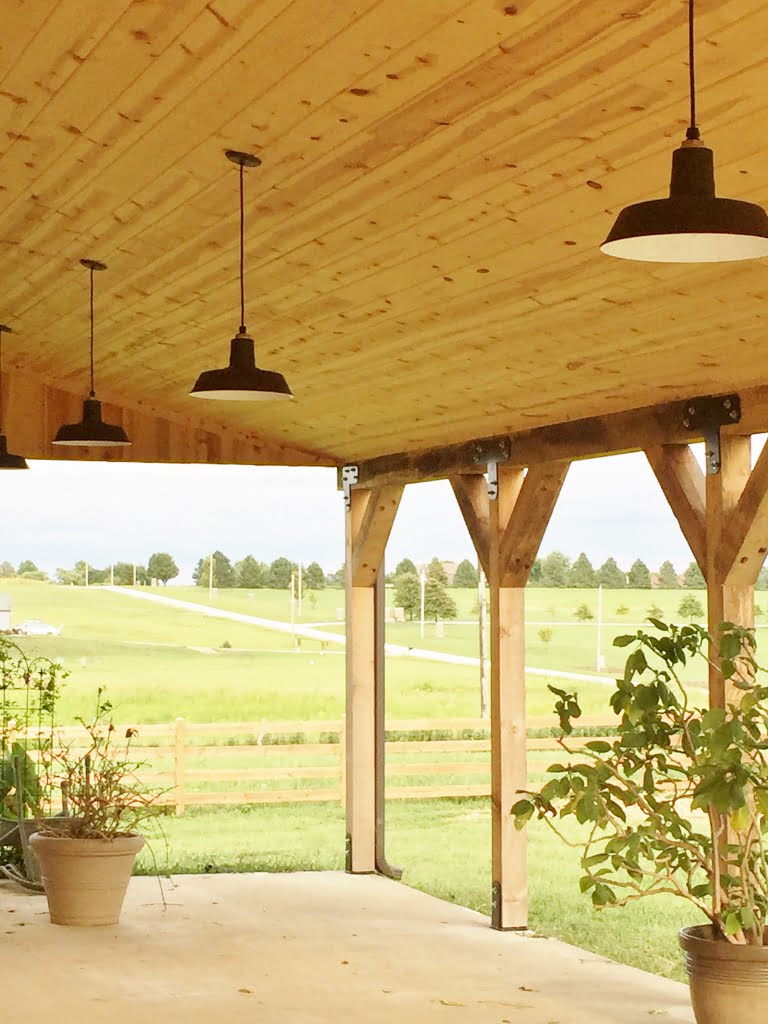 Three black barn-style pendant lights hang from a wooden ceiling on a covered porch overlooking a field