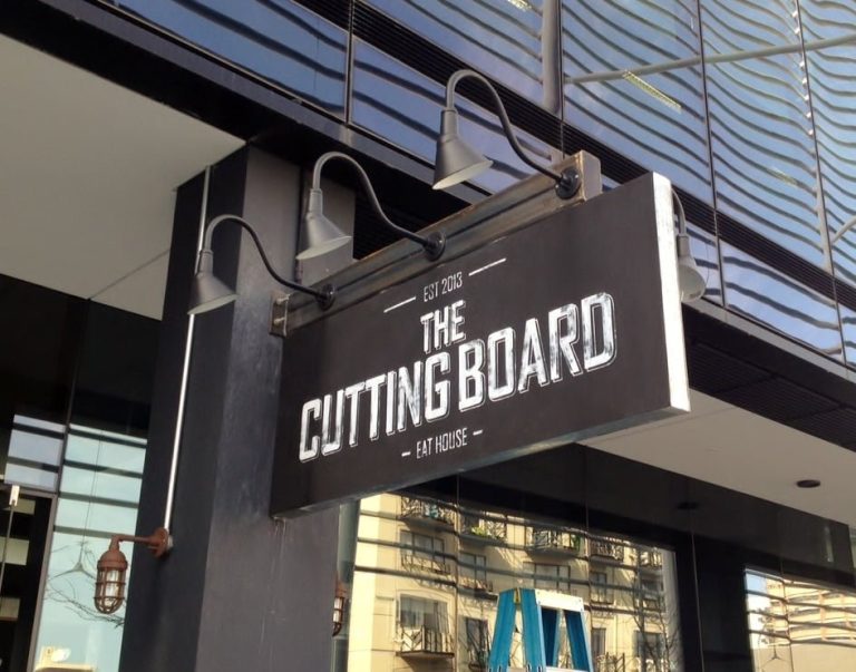 Two gooseneck barn lights illuminate a sign for 'The Cutting Board - Eat House' outside a modern building
