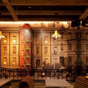 A restaurant interior features industrial-style pendant lights and a gooseneck wall light against a backdrop mural of city buildings and a world map